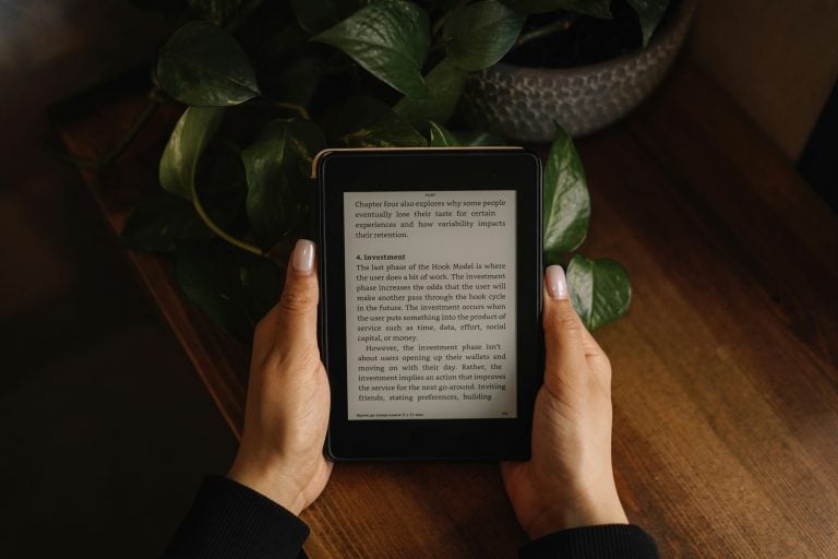 Person holding an e-reader displaying text about investment, with a green plant and wooden surface in the background.