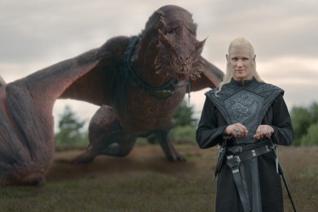 A person with long blond hair in black armor stands in front of a large, winged dragon on a grassy field.