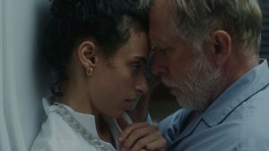 ‘Night Nurse’ (2026) Review – Seduction By Playing Helpless A woman and an older man touch foreheads closely, both appearing serious. The woman wears a white shirt and gold earring; the man has gray hair and a beard, wearing a blue shirt.