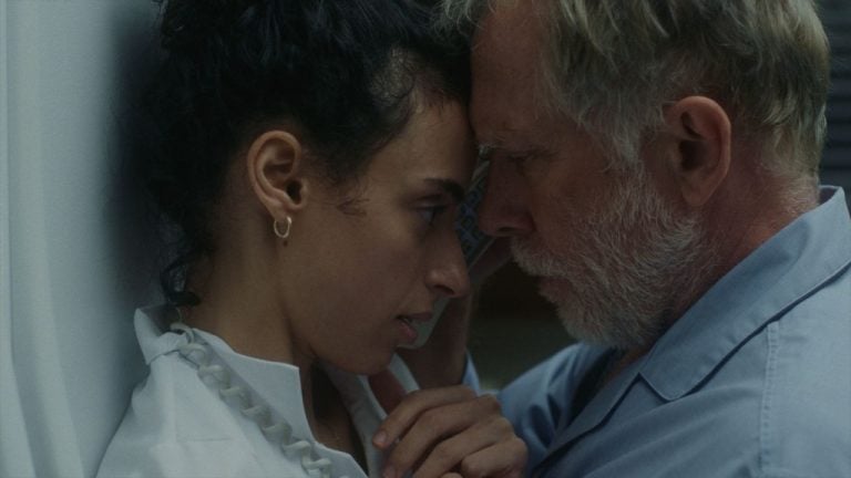 ‘Night Nurse’ (2026) Review – Seduction By Playing Helpless A woman and an older man touch foreheads closely, both appearing serious. The woman wears a white shirt and gold earring; the man has gray hair and a beard, wearing a blue shirt.
