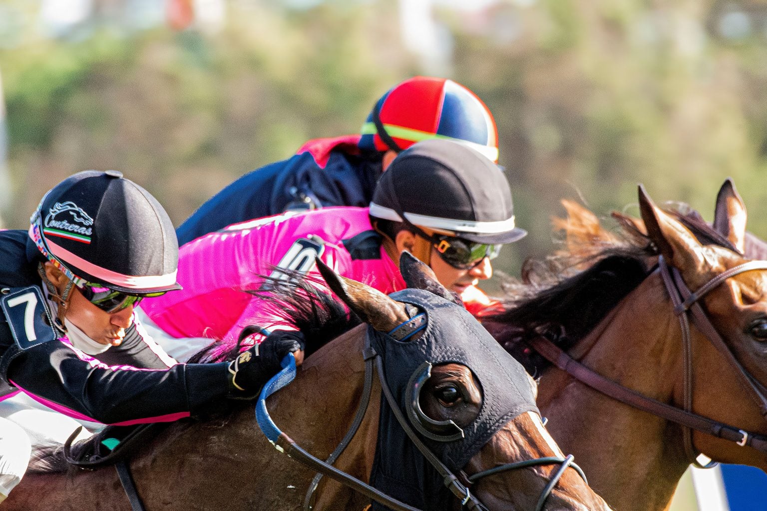2026 Haskell Stakes Betting Guide: Monmouth Park Pace And Conditions Three jockeys ride brown horses at a horse race, focused and leaning forward. The foreground jockey wears black and pink; another wears a colorful helmet behind.