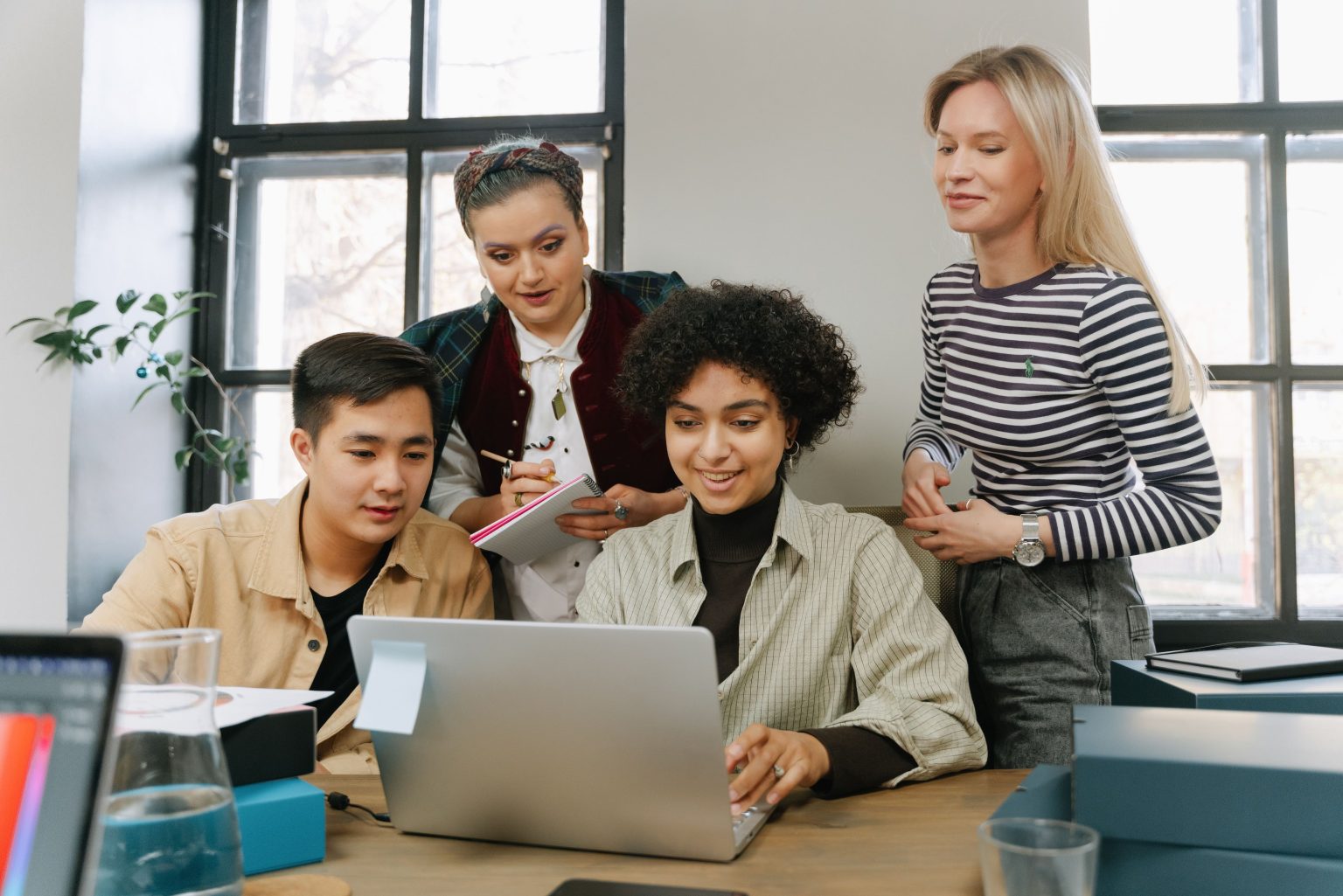 How Small Teams Can Make Tech Changes Less Stressful Four people gathered around a laptop in a bright office space, collaborating and taking notes while looking at the screen.