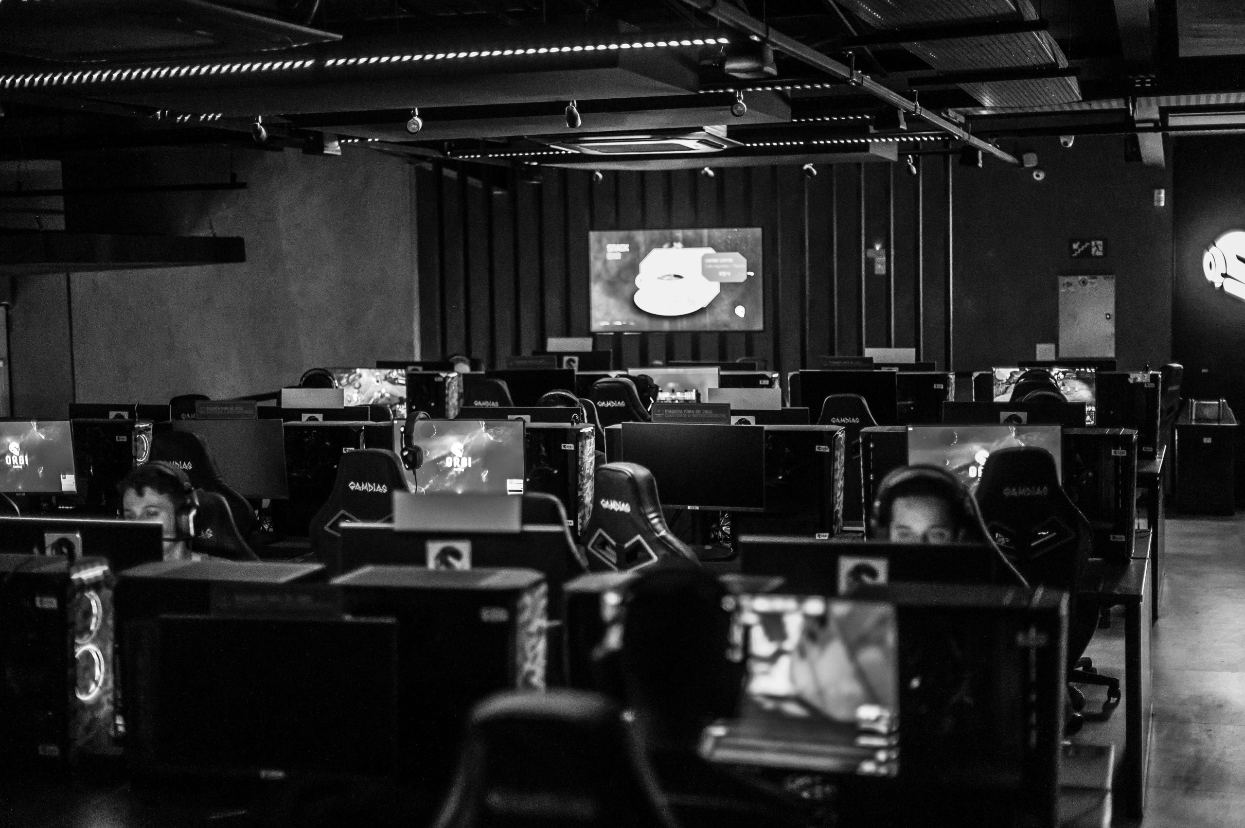 Black and white photo of a dimly lit gaming room with multiple people at computer stations and a large screen at the front.