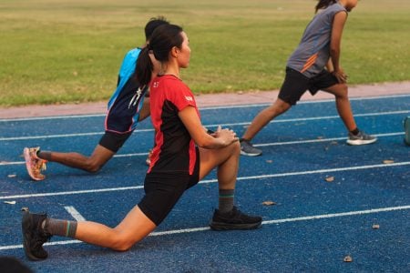 Longevity Training: Fitness Habits That Support Long-Term Performance Three people on a blue track perform lunging stretches, preparing for exercise on a sports field in the background.