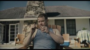 A man in a gray tank top sits in a wooden chair outdoors, holding sunglasses, with a house and stone chimney in the background.