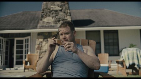 A man in a gray tank top sits in a wooden chair outdoors, holding sunglasses, with a house and stone chimney in the background.