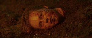 A severed human head with blood and dirt on the face lies partially buried in soil, eyes open, under dim, reddish light.