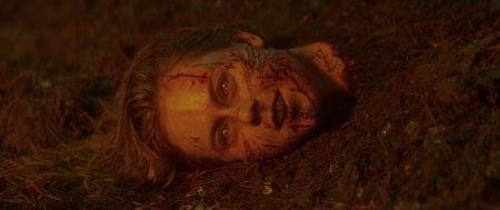 A severed human head with blood and dirt on the face lies partially buried in soil, eyes open, under dim, reddish light.