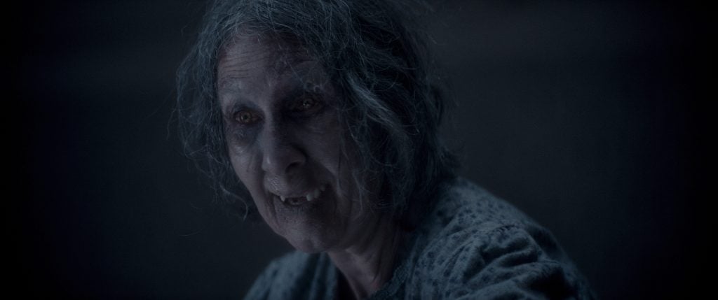 An elderly woman with disheveled gray hair, sunken eyes, and decayed teeth stares ahead in dim lighting, appearing distressed or unwell.