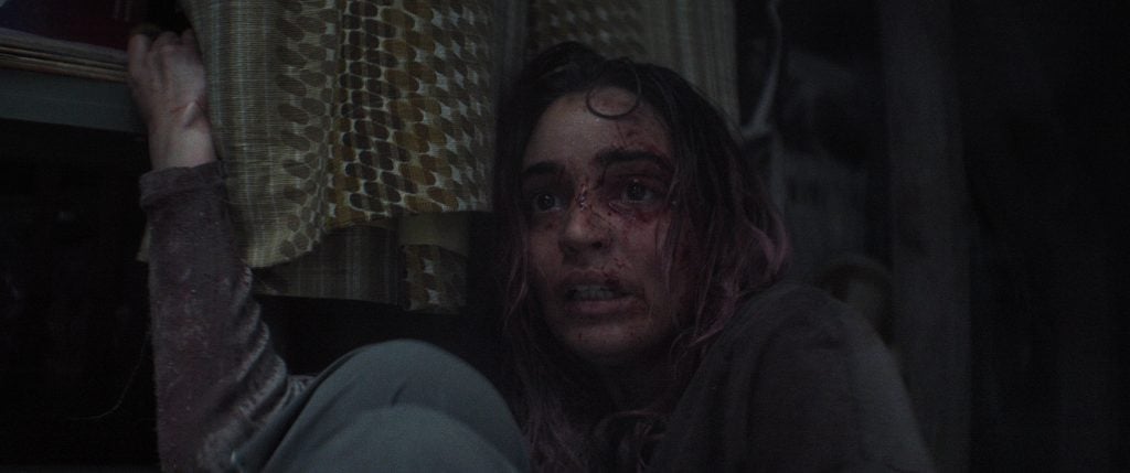 A woman with blood and dirt on her face sits against a wall, looking up with a fearful expression. Yellow patterned curtains are visible beside her.