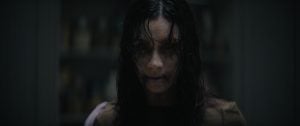 A person with long dark hair, pale skin, and dark circles under their eyes stares forward with a menacing expression in a dimly lit room.