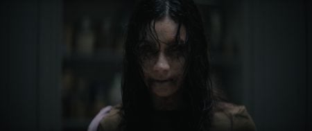 A person with long dark hair, pale skin, and dark circles under their eyes stares forward with a menacing expression in a dimly lit room.