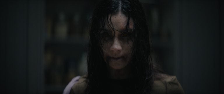 ‘Evil Dead Burn’ Review – Another Brutally Bloody And Nasty Entry In A Franchise That Consistently Delivers The Goods A person with long dark hair, pale skin, and dark circles under their eyes stares forward with a menacing expression in a dimly lit room.