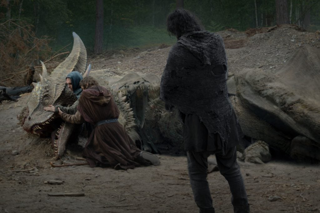 Two people kneel beside a large dragon lying on the ground in a forest, gently touching its head, while a third person in dark clothing stands nearby watching.