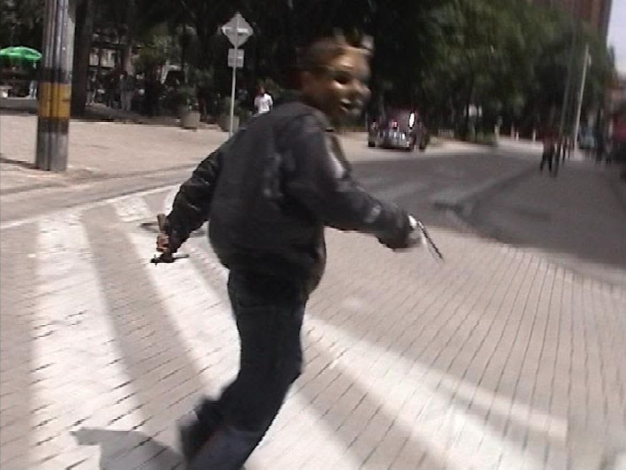A person wearing a mask and holding an object stands on a crosswalk in an urban area, looking back toward the camera.