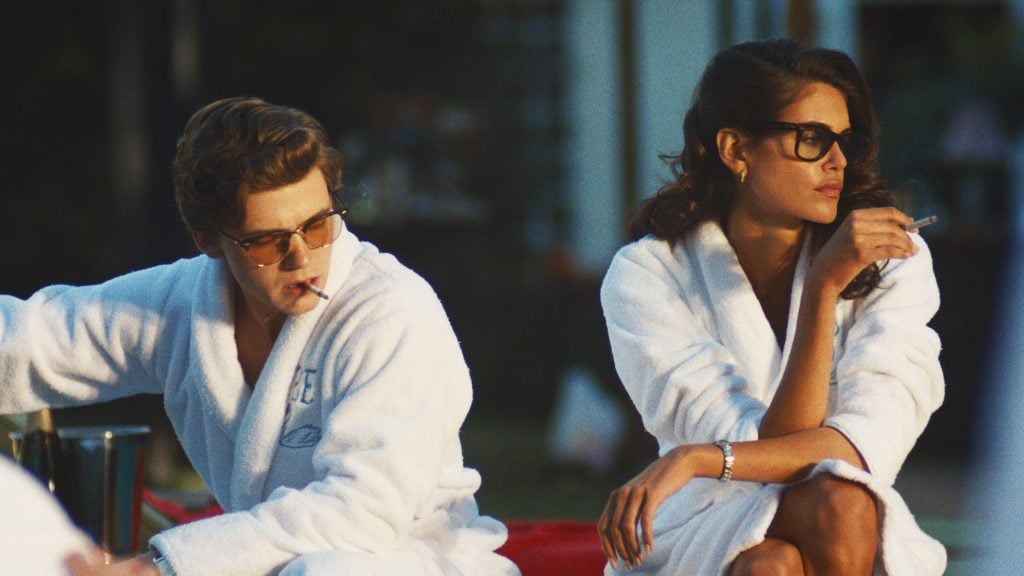 Two people wearing white robes and glasses sit outdoors, each holding a cigarette, looking in opposite directions.