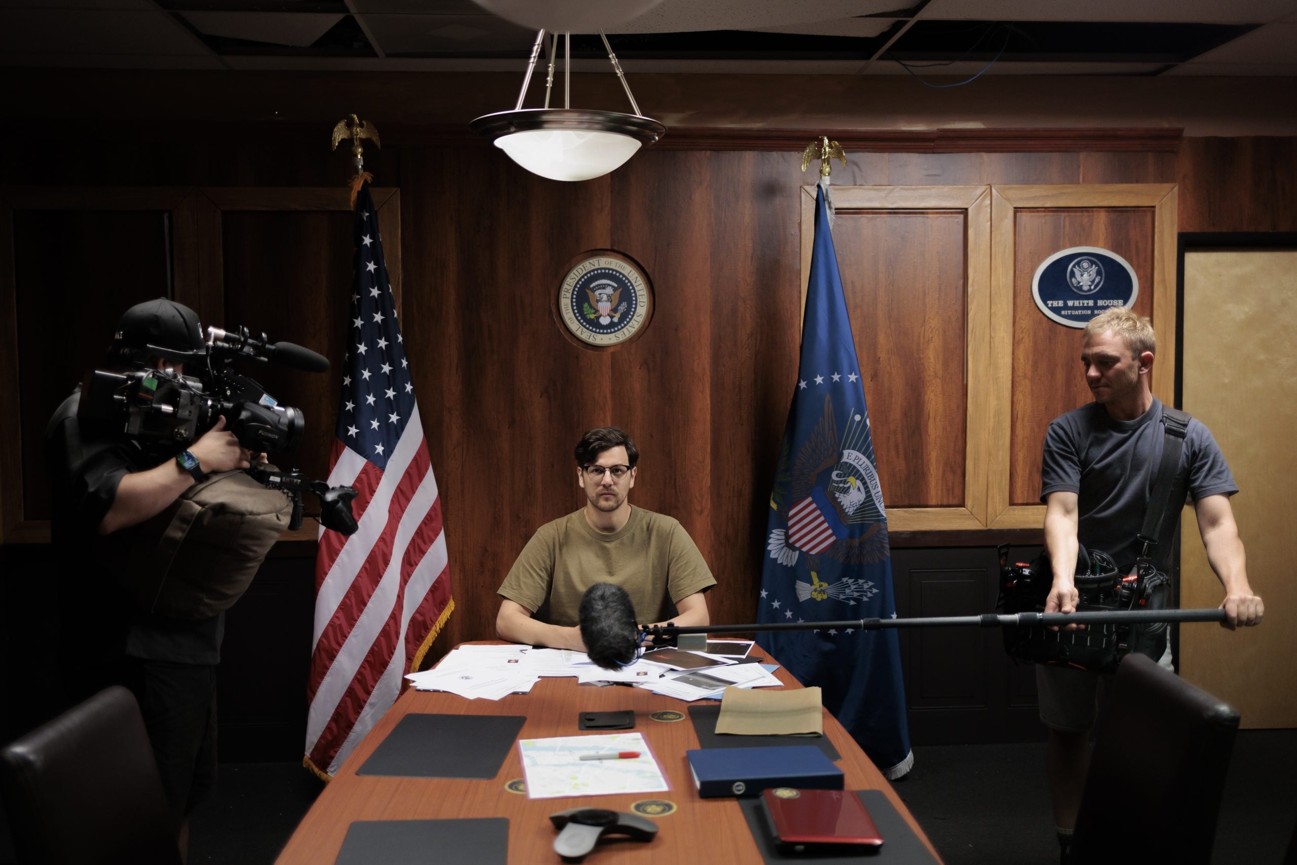 A man sits at a desk with papers in a room decorated with U.S. flags and presidential seals, while two crew members operate a camera and boom microphone.