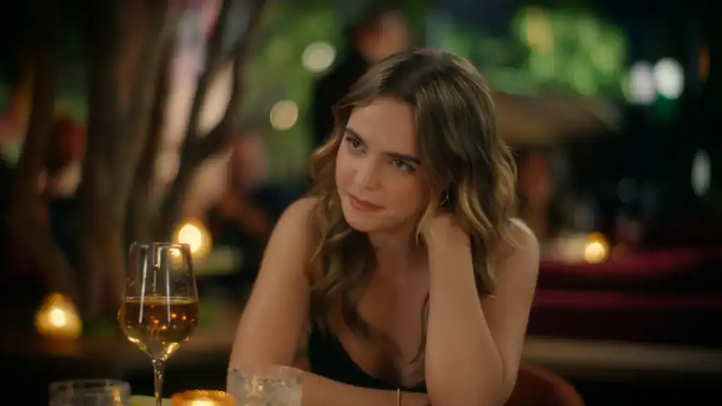 A woman with long wavy hair sits at a dimly lit table, resting her head on her hand, with a wine glass and candles in front of her.