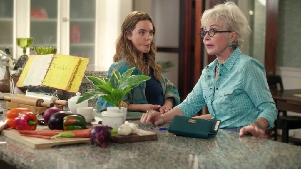 Two women sit at a kitchen counter with vegetables, a cookbook, and a purse in front of them. One looks forward while the other gazes to the side.