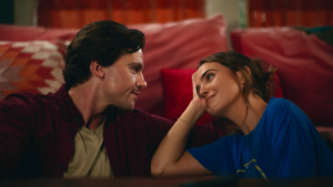 A man and a woman sit close together on a couch, facing each other and smiling, with red cushions in the background.