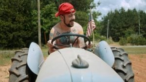 A man wearing a red cap sits shirtless on a tractor, smiling and holding the steering wheel, with an American flag visible behind him.