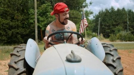 A man wearing a red cap sits shirtless on a tractor, smiling and holding the steering wheel, with an American flag visible behind him.