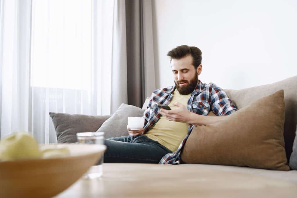 Man sitting on a sofa, holding a cup in one hand and looking at his smartphone in the other, with neutral-colored pillows and a bowl of fruit in the foreground.