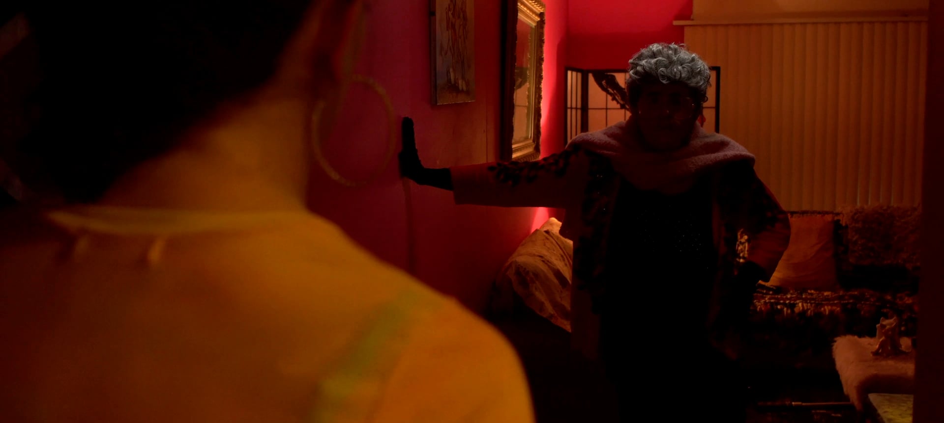 A person with gray hair and glasses stands in a dimly lit hallway facing another person wearing large hoop earrings, with both partially illuminated by warm lighting.