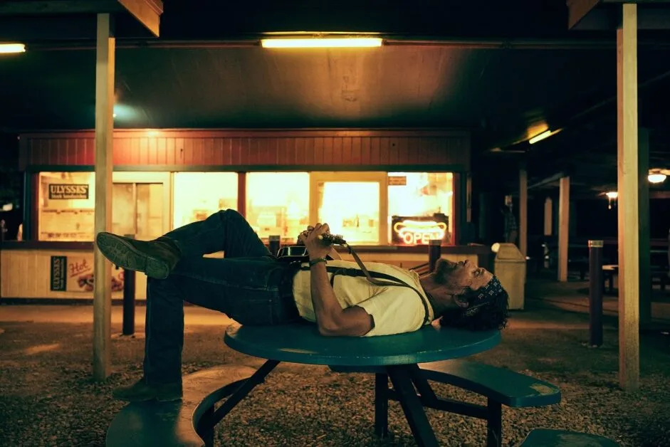 A man lies on his back atop a blue picnic table at night, holding a guitar, with a lit “OPEN” sign in the background.