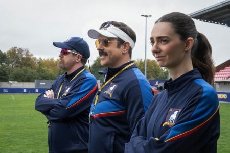 ‘Ted Lasso’ Season 4 Review – He Shoots, He Misses Three coaches wearing navy tracksuits stand side by side on a soccer field, looking ahead with arms crossed. Stadium seating and trees are visible in the background.