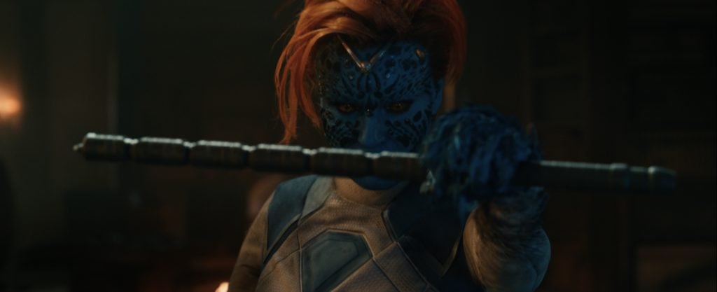 A blue-skinned character with orange hair and patterned face holds a staff, extending it forward in a dimly lit room.