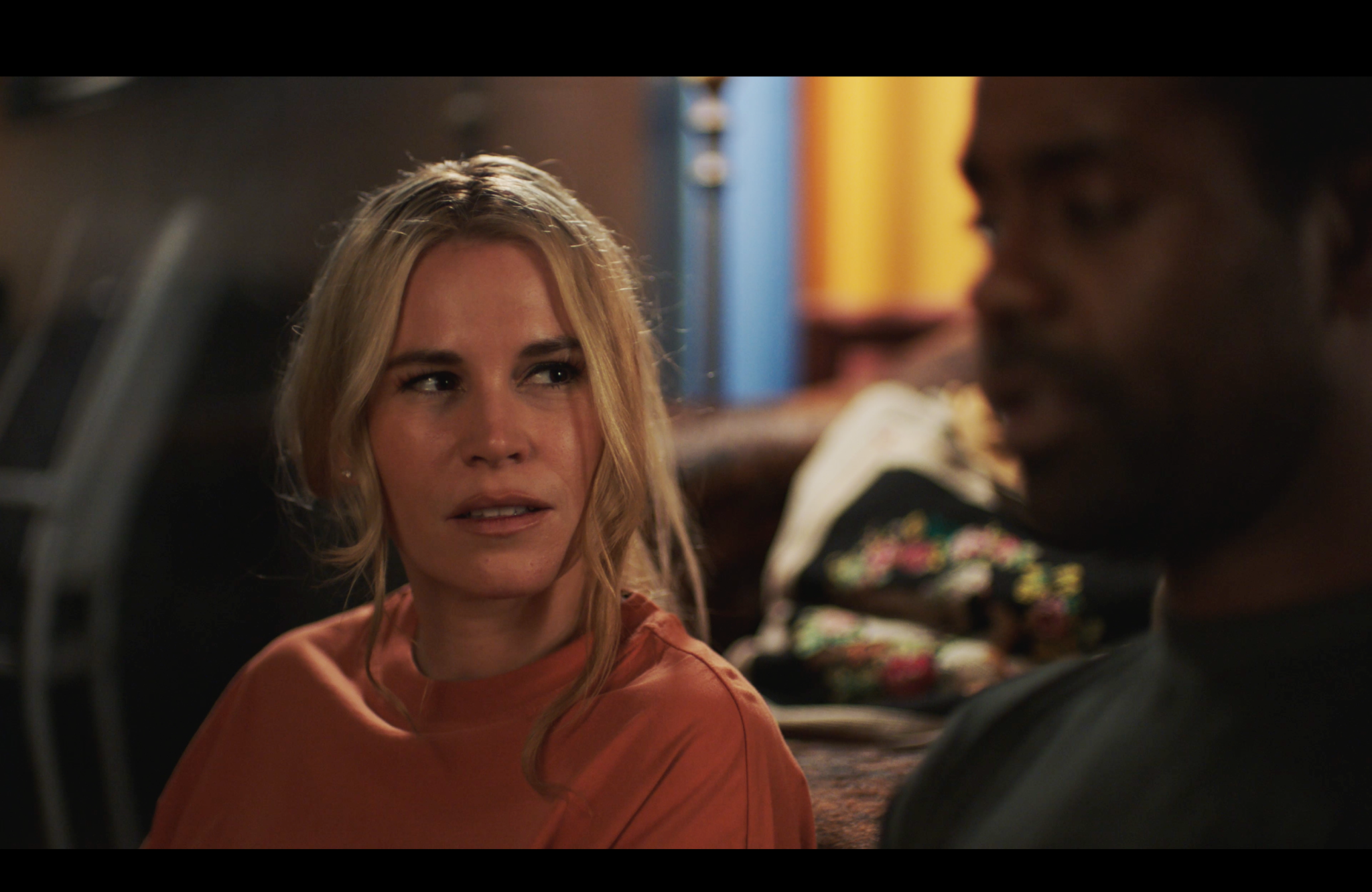 A woman with blond hair in an orange shirt sits indoors, looking at a man next to her with a serious expression. The background is softly blurred.
