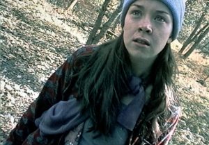 A young woman wearing a blue beanie and scarf stands in a wooded area, looking ahead with a concerned expression. The image has a slightly tilted angle and a grainy quality.