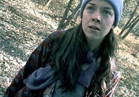 Lionsgate To Give ‘The Blair Witch Project’ A Collector’s Edition Blu-Ray Release This October A young woman wearing a blue beanie and scarf stands in a wooded area, looking ahead with a concerned expression. The image has a slightly tilted angle and a grainy quality.