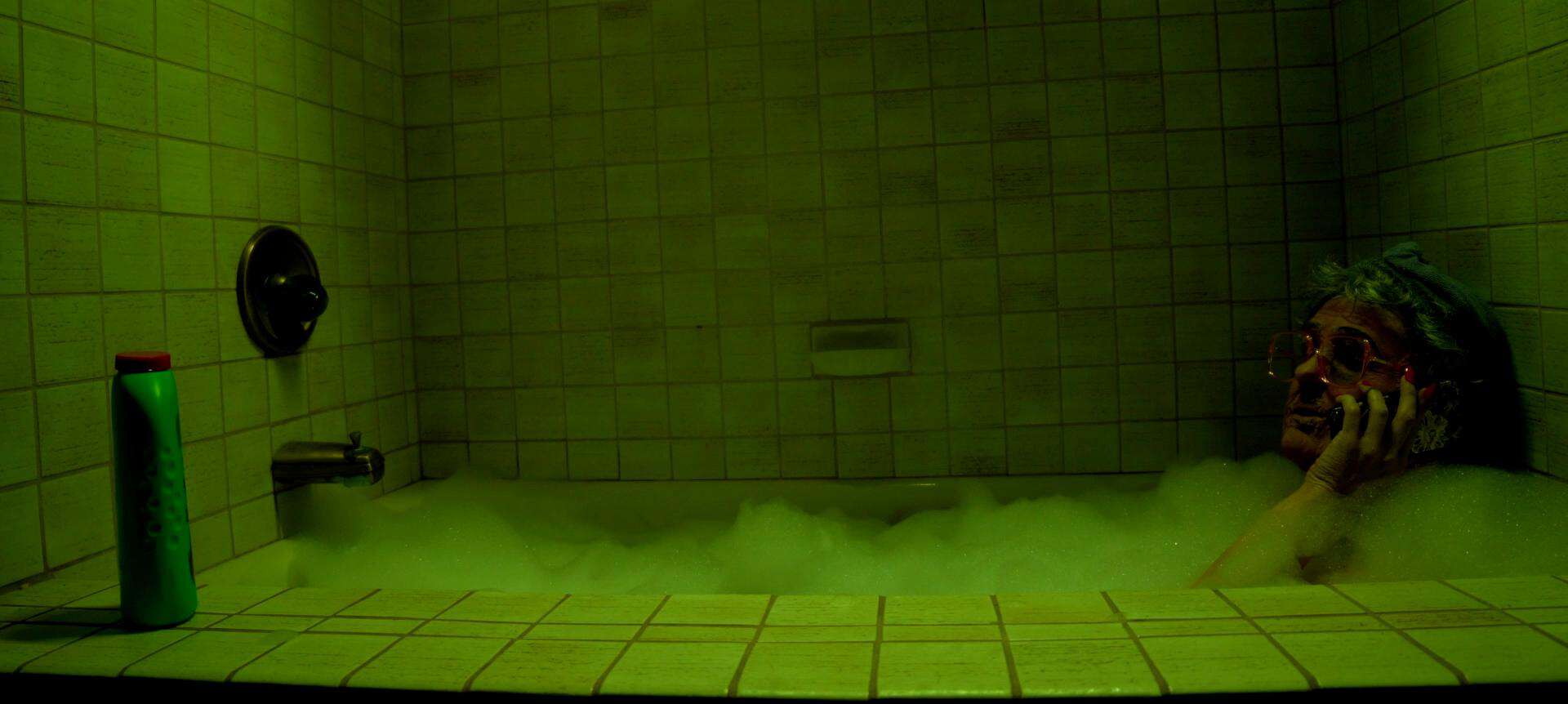 Person wearing sunglasses and a towel on their head sits in a bubble-filled bathtub, talking on the phone. A green bottle is placed on the bathtub ledge. The lighting is dim and greenish.