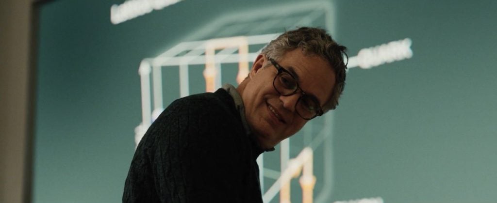 A man with glasses and gray hair stands in front of a projection of a diagram on a green screen, looking over his shoulder and smiling.