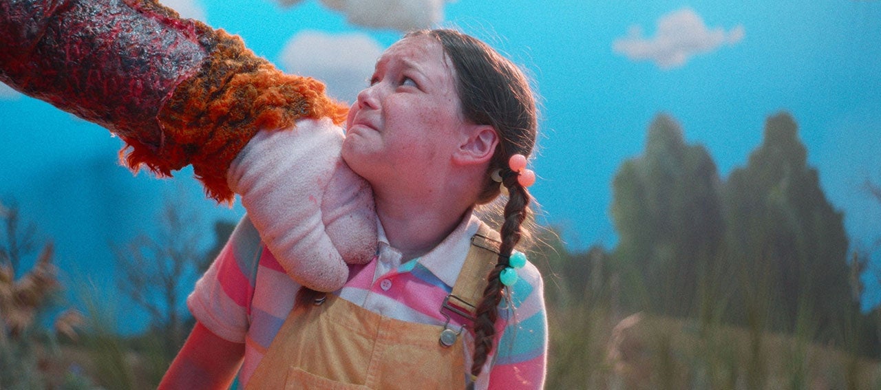 A person in a furry costume grips a young girl by the neck outdoors. The girl looks scared and wears yellow overalls and pink shirt. The background shows a blue sky and trees.