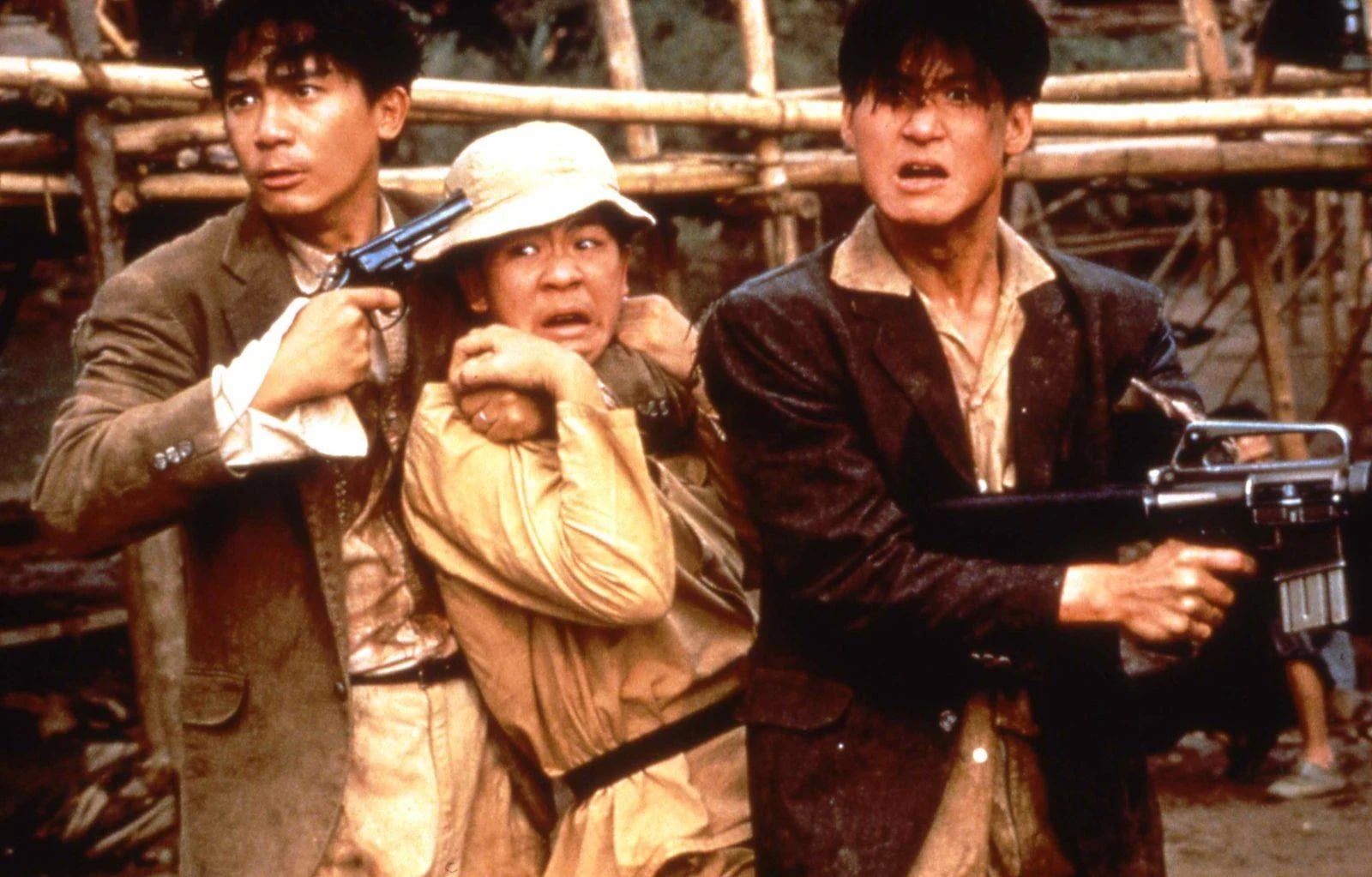 Three men outdoors, one holding another hostage with a gun to his head, while the third man points a large firearm at them. Wooden scaffolding is visible in the background.