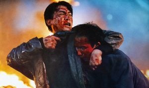 Two bloodied men stand amid flames; one holds a gun to his own head while restraining the other, both appearing distressed in a tense, dramatic scene.