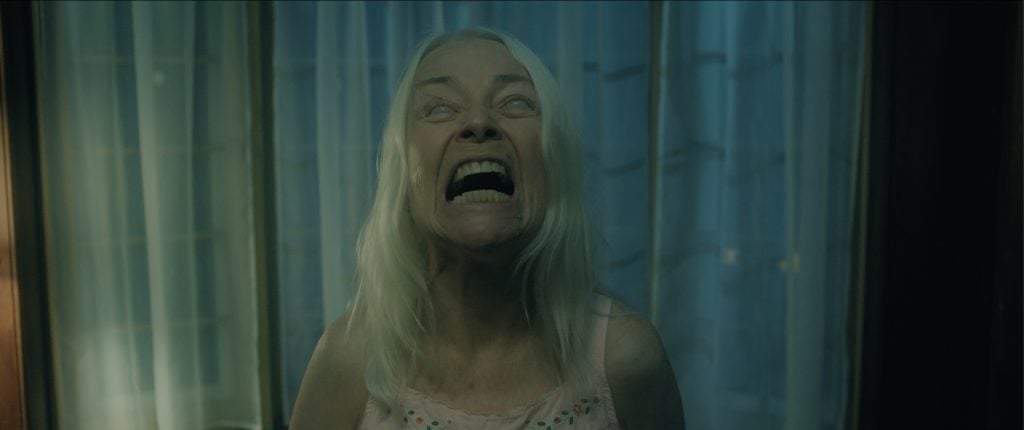 An older woman with long white hair stands in front of curtains, tilting her head back with her eyes rolled upward and mouth wide open in a scream.