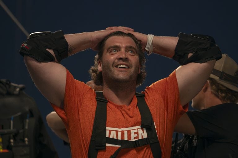 The Challenge 42 Cutthroat Interview – Team Orange Talks Cheating Scandal The Challenge A man in an orange shirt with the word “CHALLENGE” stands with his hands on his head, smiling, wearing black padded gear, and surrounded by people.