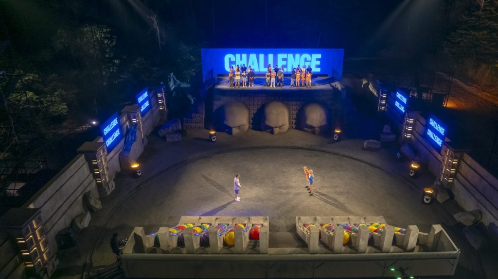 Wide shot of a stage set for a competition show, with two contestants in the center and a large “CHALLENGE” sign on the back wall.