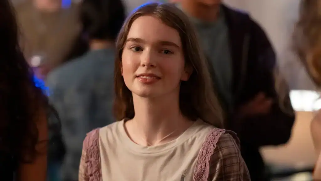 A young woman with long brown hair stands indoors, looking at the camera and smiling slightly, with blurred people in the background.