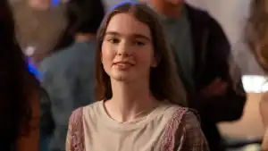 A young woman with long brown hair stands indoors, looking at the camera and smiling slightly, with blurred people in the background.