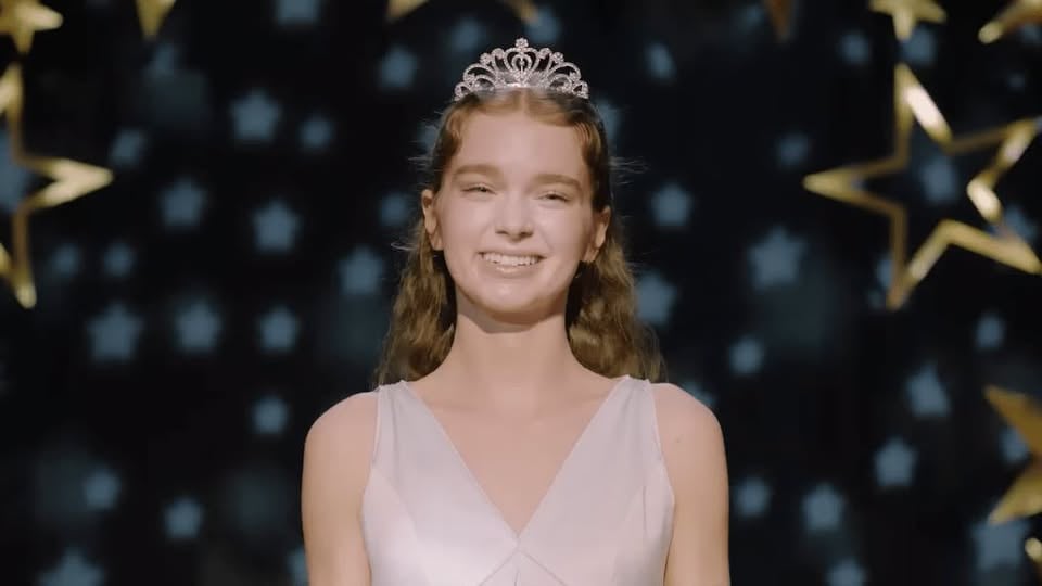 A young person wearing a tiara and a light dress smiles while standing in front of a backdrop decorated with stars.