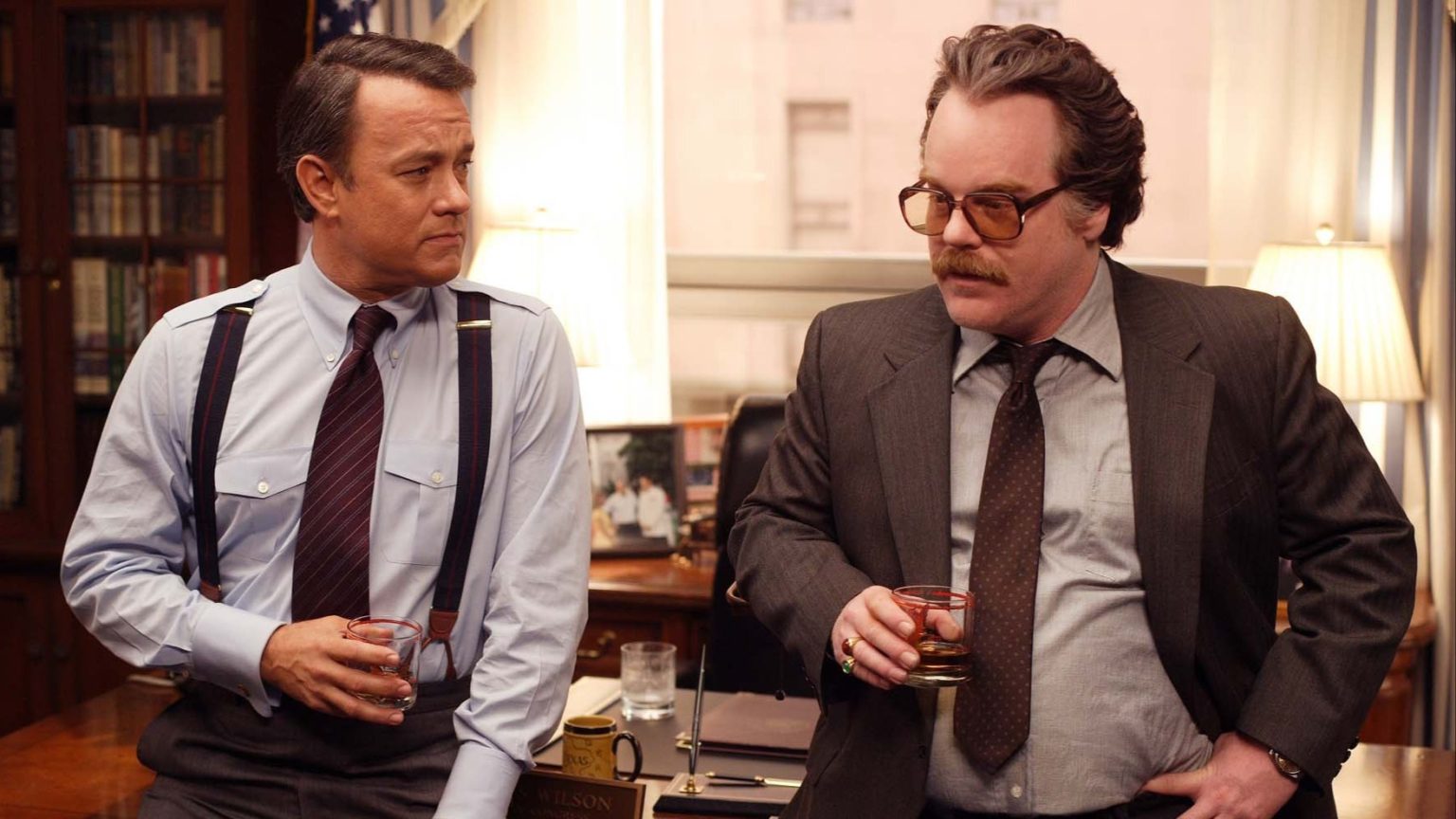 Two men in suits stand in an office, each holding a glass of whiskey and engaged in conversation. One man wears suspenders, the other glasses. Bookshelves and lamps are visible in the background.