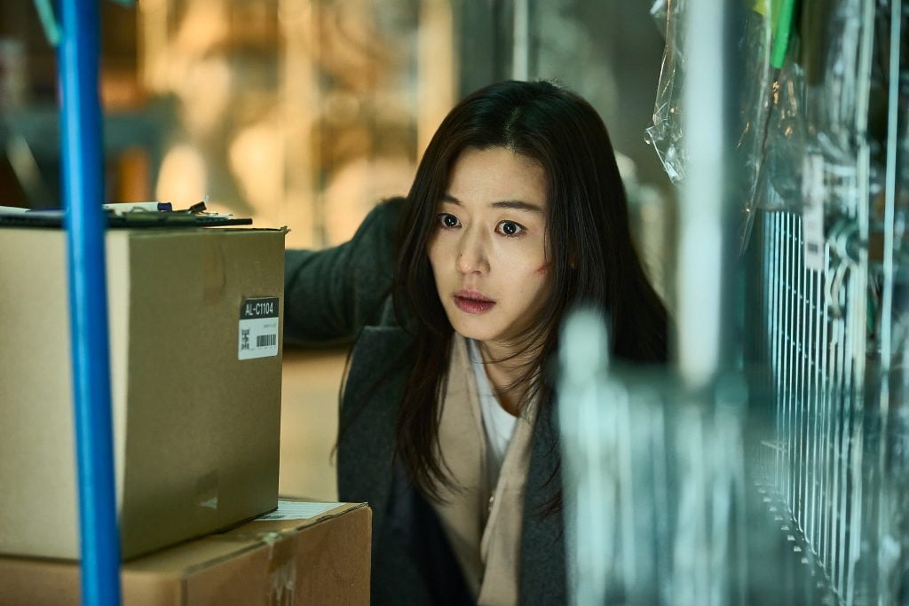 A woman with long dark hair crouches behind cardboard boxes in a storage area, looking alert and concerned.