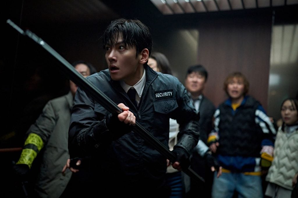 A security guard holds a long weapon and stands protectively in front of a group of frightened people inside an elevator.