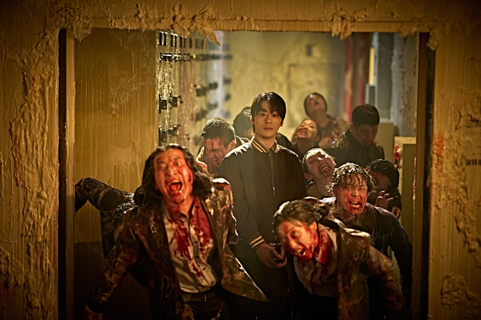 ‘Colony’ (2026) Review – Yeon Sang-ho’s Return to Zombie Flicks Never Lets Up On the Carnage A young man stands calmly in a doorway as several bloodied, zombie-like figures lunge around him, creating a chaotic scene in a dimly lit hall.