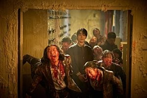‘Colony’ (2026) Review – Yeon Sang-ho’s Return to Zombie Flicks Never Lets Up On the Carnage A young man stands calmly in a doorway as several bloodied, zombie-like figures lunge around him, creating a chaotic scene in a dimly lit hall.